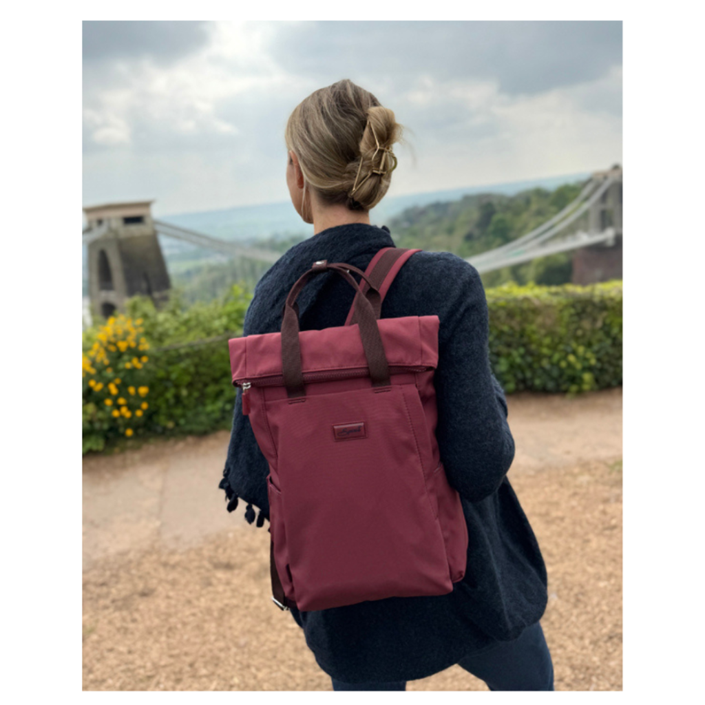 A woman seen from behind wearing a burgundy roll-top backpack with dark brown handles, standing outdoors in front of a suspension bridge and greenery.
