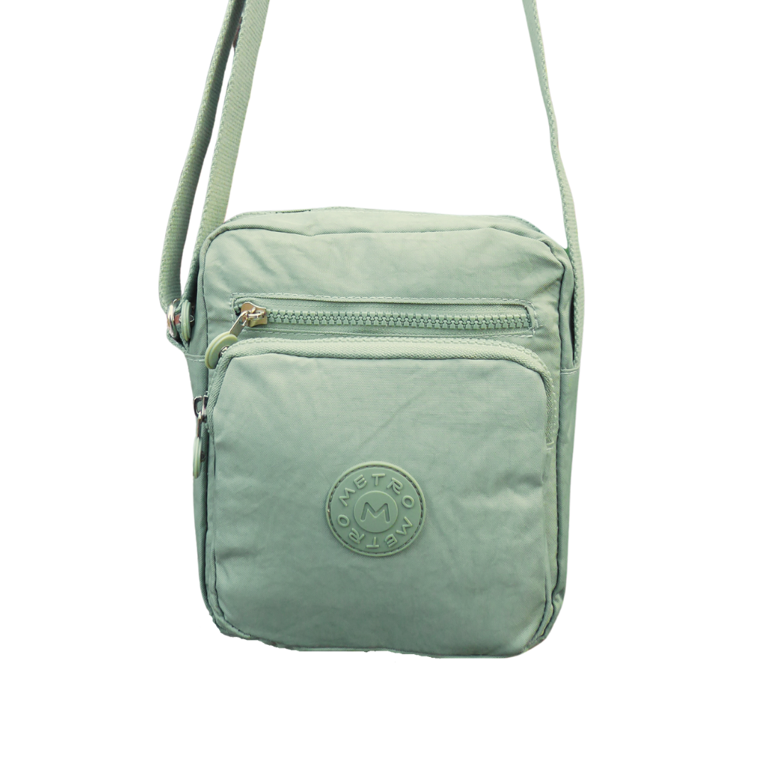 A small crossbody bag in light green, featuring a front zippered pocket with a  logo badge, a top zippered compartment, and a matching shoulder strap, shown against a plain white background.