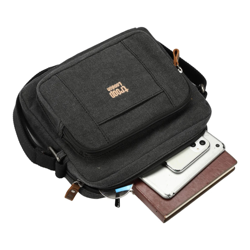 A black canvas Troop London Small Canvas Cross-body Bag - Lightweight & Brass Hardware with a zippered front pocket and brown leather accents, shown with a smartphone and notebook partially tucked into its open side pocket on a white background.